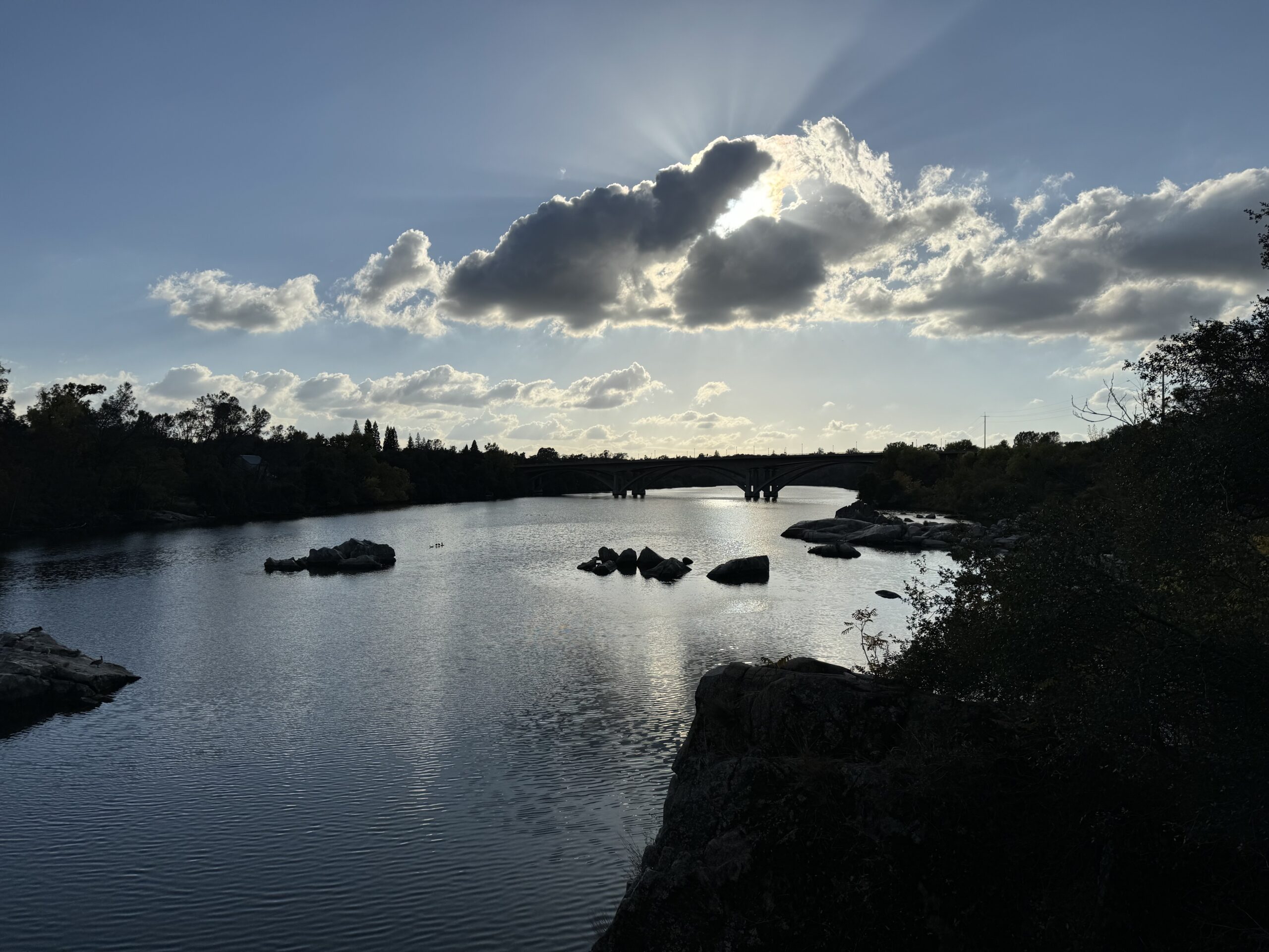 A lake with rocks and trees on the side in shadow. There is a bridge over the lake, and the sun is shining out in bursting rays from behind a cloud.