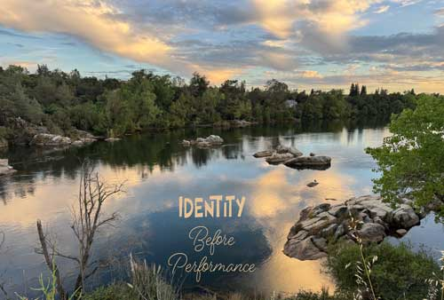 A peaceful river at sunset reflects golden clouds and a soft blue sky, with trees and large granite rocks lining the shoreline. The calm water mirrors the light above, creating a tranquil scene that evokes reflection and rest. Overlaid text reads: "Identity Before Performance."